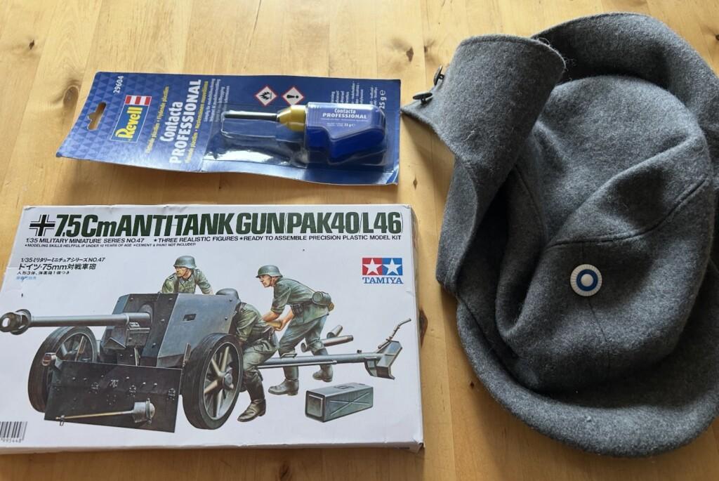 Kuvassa pienoismalli, liimatuubi ja villakankainen harmaa hattu kokardeineen.
